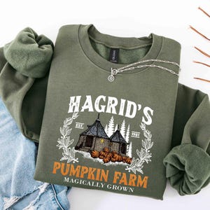 May include: An olive green sweatshirt with the text "Hagrid's Pumpkin Farm" in white and orange. The design includes an illustration of a house, pumpkins, and the text "Magically Grown". A silver necklace is draped across the sweatshirt.