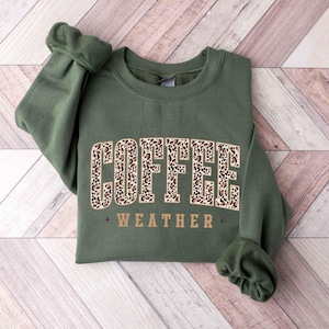 May include: An olive green sweatshirt with the words "COFFEE WEATHER" in a brown leopard print design. The sweatshirt is folded on a light brown wooden surface.