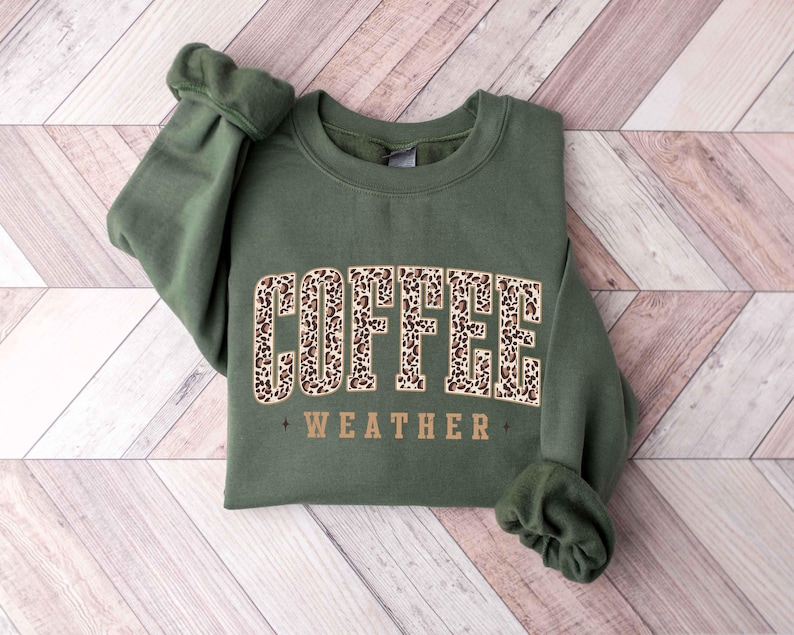 May include: An olive green sweatshirt with the words "COFFEE WEATHER" in a brown leopard print design. The sweatshirt is folded on a light brown wooden surface.