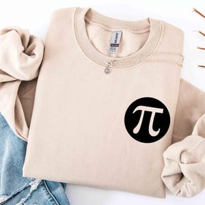 May include: A beige crewneck sweatshirt with a black circle graphic featuring the Greek letter pi. A silver necklace with a sunburst pendant is draped across the folded sweatshirt. The sweatshirt is folded on top of a pair of blue jeans.