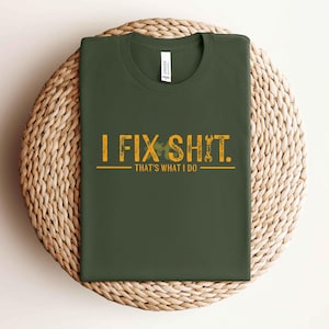 May include: Dark green t-shirt with the text "I FIX SHIT. That's what I do" in distressed yellow lettering. The design includes wrench and shamrock graphics. The shirt is folded on a woven, circular surface.