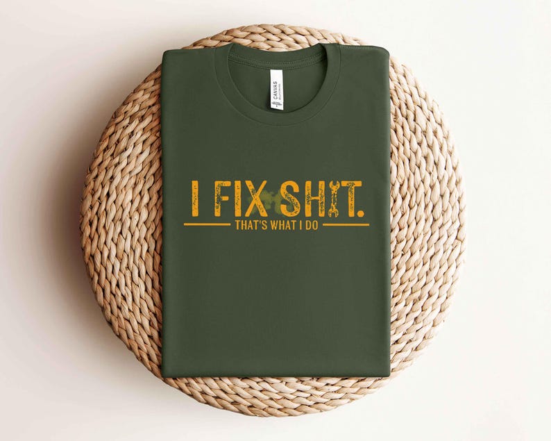 May include: Dark green t-shirt with the text "I FIX SHIT. That's what I do" in distressed yellow lettering. The design includes wrench and shamrock graphics. The shirt is folded on a woven, circular surface.