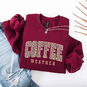 May include: A maroon crewneck sweatshirt with the words "COFFEE WEATHER" in a leopard print design. A silver necklace with a pendant is on the sweatshirt. Light blue ripped jeans are visible on the left side of the image.