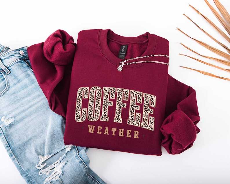 May include: A maroon crewneck sweatshirt with the words "COFFEE WEATHER" in a leopard print design. A silver necklace with a pendant is on the sweatshirt. Light blue ripped jeans are visible on the left side of the image.