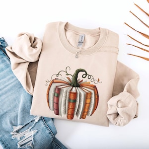 May include: Beige sweatshirt featuring a pumpkin design made of books. The pumpkin has a green stem and vines. The books are in shades of orange, red, and brown. The sweatshirt is paired with blue jeans.