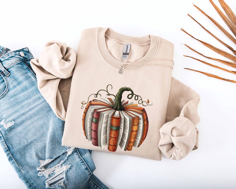 May include: Beige sweatshirt featuring a pumpkin design made of books. The pumpkin has a green stem and vines. The books are in shades of orange, red, and brown. The sweatshirt is paired with blue jeans.
