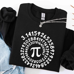 May include: Black sweatshirt with a white Pi symbol and the digits of Pi arranged in a circle. A silver necklace with a small pendant is draped over the sweatshirt. The sweatshirt is folded on top of blue jeans.