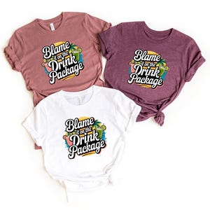May include: Three t-shirts in shades of mauve, purple, and white, each featuring the phrase "Blame it on the Drink Package" in a playful font. The graphic includes cocktail and palm tree illustrations, suggesting a vacation or party theme.