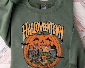 Può includere: Una felpa girocollo verde oliva con una grande grafica di una zucca arancione. La zucca contiene una scena dettagliata di una città con edifici e una zucca più piccola. Il testo recita HALLOWEENTOWN e EST. 1998 con pipistrelli neri.