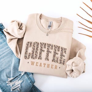 May include: A beige sweatshirt with the words "COFFEE WEATHER" in a leopard print design. The sweatshirt is folded and placed on a white surface next to a pair of blue jeans.