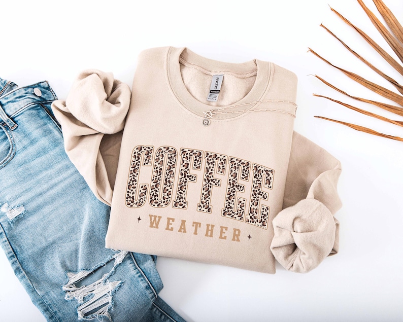 May include: A beige sweatshirt with the words "COFFEE WEATHER" in a leopard print design. The sweatshirt is folded and placed on a white surface next to a pair of blue jeans.