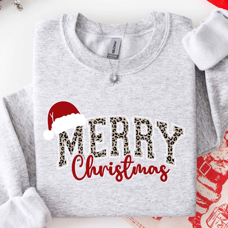 May include: A light gray sweatshirt with the words "MERRY Christmas" in a festive design. The word "MERRY" has a leopard print pattern, and a red Santa hat is above it. The word "Christmas" is in red script.