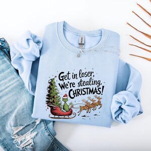May include: Light blue sweatshirt with the text "Get in loser, We're stealing CHRISTMAS!". The graphic features a green character in a Santa hat on a red sleigh with a Christmas tree and reindeer. The sweatshirt is paired with blue jeans.