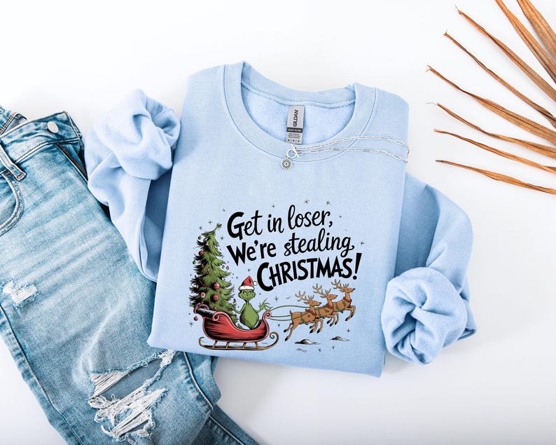 May include: Light blue sweatshirt with the text "Get in loser, We're stealing CHRISTMAS!". The graphic features a green character in a Santa hat on a red sleigh with a Christmas tree and reindeer. The sweatshirt is paired with blue jeans.