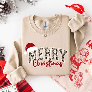 May include: Beige crewneck sweatshirt with "MERRY Christmas" text. The word "MERRY" has a leopard print design, and a Santa hat is above it. A red "Christmas" is below. A Christmas-themed gift bag and decorations are in the background.