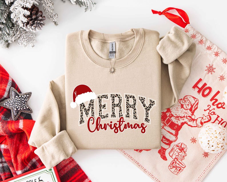 May include: Beige crewneck sweatshirt with "MERRY Christmas" text. The word "MERRY" has a leopard print design, and a Santa hat is above it. A red "Christmas" is below. A Christmas-themed gift bag and decorations are in the background.