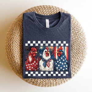 May include: A navy blue t-shirt featuring a graphic of three chickens with patriotic designs. The chickens are decorated with red, white, and blue stars and stripes, and are set against a checkered border.
