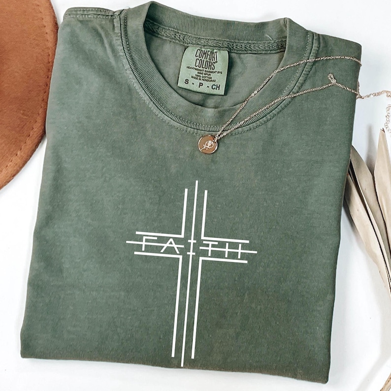 May include: An olive green t-shirt with the word "FAITH" in white, designed as a cross. A gold-colored necklace with a small pendant rests on the shirt. The shirt has a Comfort Colors label, size S-P-CH.