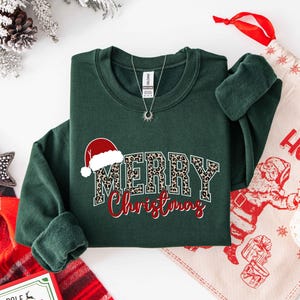May include: A dark green sweatshirt with the words "MERRY Christmas" in a festive design. The word "MERRY" has a leopard print pattern, and the word "Christmas" is in red script. A Santa hat graphic is above the word "MERRY".