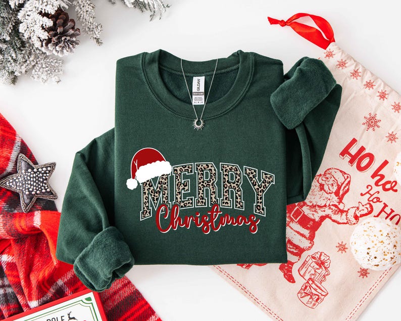 May include: A dark green sweatshirt with the words "MERRY Christmas" in a festive design. The word "MERRY" has a leopard print pattern, and the word "Christmas" is in red script. A Santa hat graphic is above the word "MERRY".