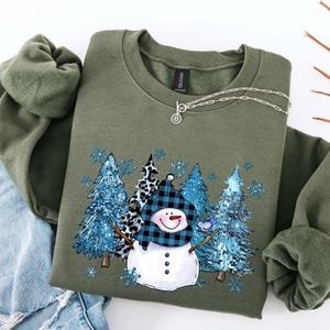 May include: An olive green sweatshirt featuring a snowman graphic with a blue and black plaid scarf and hat. The design includes blue glittery trees and snowflakes. A silver necklace with a sun pendant is draped on the sweatshirt.