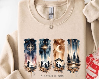 May include: Beige sweatshirt with a watercolor design featuring a star, moon, and silhouettes of figures. The design includes the text "A Savior Is Born". The sweatshirt has a crew neck and long sleeves.