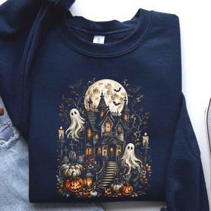 May include: A navy blue crewneck sweatshirt featuring a detailed graphic print of a haunted house under a full moon. The design includes two white ghosts, orange pumpkins, lit candles, gravestones, and flying bats in a dark, atmospheric scene.