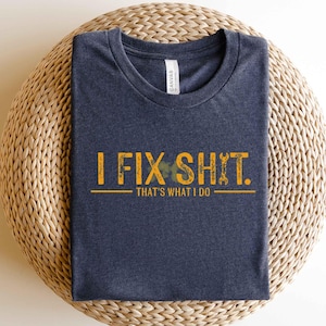 May include: Navy blue t-shirt with the text "I FIX SHIT. THAT'S WHAT I DO" in yellow. The design includes a wrench graphic. The shirt is folded on a woven, circular surface.