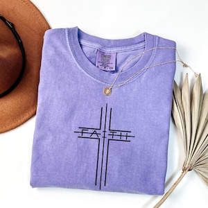 May include: A lavender Comfort Colors t-shirt with a black cross design and the word "Faith" printed across the center. A gold necklace with a pendant is draped over the shirt. A brown hat and dried palm leaf are in the background.