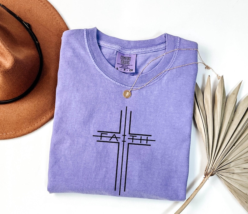 May include: A lavender Comfort Colors t-shirt with a black cross design and the word "Faith" printed across the center. A gold necklace with a pendant is draped over the shirt. A brown hat and dried palm leaf are in the background.