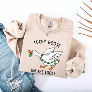 May include: Beige sweatshirt with the text "LUCKY GOOSE ON THE LOOSE" and a cartoon goose design. The goose has a green shamrock garland and is holding a shamrock in its beak. The sweatshirt is folded next to a pair of blue jeans.
