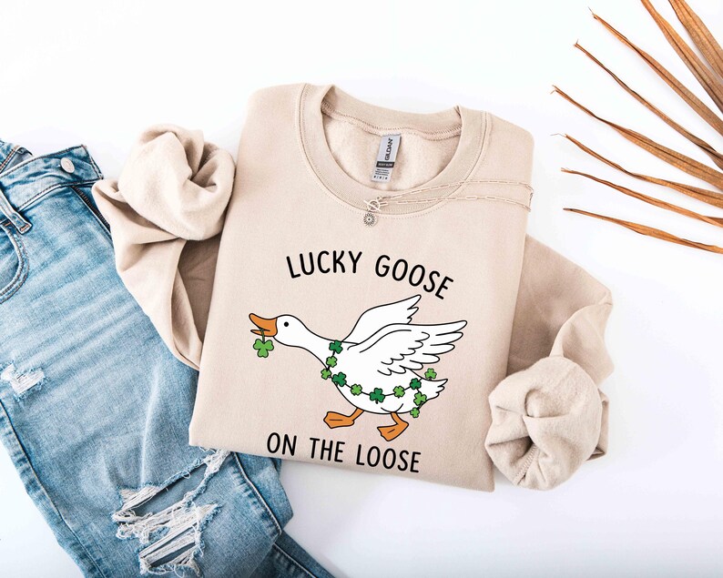 May include: Beige sweatshirt with the text "LUCKY GOOSE ON THE LOOSE" and a cartoon goose design. The goose has a green shamrock garland and is holding a shamrock in its beak. The sweatshirt is folded next to a pair of blue jeans.