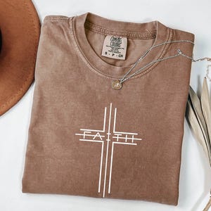 May include: A brown t-shirt with a white cross design and the word "FAITH" in the center. The shirt has a crew neck and short sleeves. A silver necklace with a small pendant rests on the shirt. The shirt is made of a soft, comfortable material.