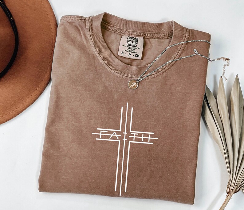 May include: A brown t-shirt with a white cross design and the word "FAITH" in the center. The shirt has a crew neck and short sleeves. A silver necklace with a small pendant rests on the shirt. The shirt is made of a soft, comfortable material.