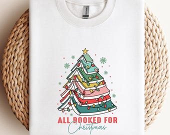 May include: White sweatshirt featuring a colorful Christmas tree design made of books, with a star on top and string lights. The text "ALL BOOKED FOR Christmas" is printed below the tree. A festive holiday-themed apparel item.
