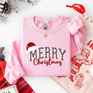 May include: A light pink sweatshirt with the words "MERRY Christmas" in a leopard print font and red script. A Santa hat graphic is above the word "MERRY". A Christmas-themed gift bag and decorations are in the background.