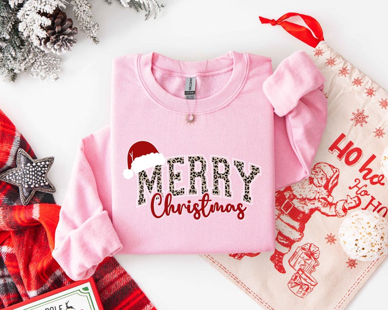 May include: A light pink sweatshirt with the words "MERRY Christmas" in a leopard print font and red script. A Santa hat graphic is above the word "MERRY". A Christmas-themed gift bag and decorations are in the background.