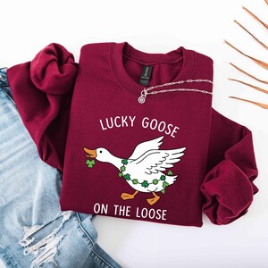 May include: A maroon sweatshirt with the text "LUCKY GOOSE ON THE LOOSE" and a graphic of a white goose wearing a shamrock garland. A silver necklace is draped on the sweatshirt. Light blue jeans are visible in the background.