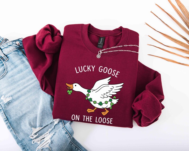 May include: A maroon sweatshirt with the text "LUCKY GOOSE ON THE LOOSE" and a graphic of a white goose wearing a shamrock garland. A silver necklace is draped on the sweatshirt. Light blue jeans are visible in the background.