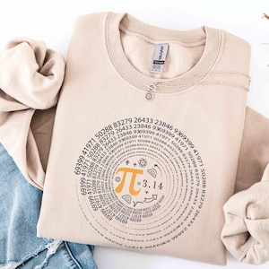 May include: Beige crewneck sweatshirt featuring a Pi symbol design with the number 3.14 and mathematical symbols. The design is in black and orange, with a circular pattern of numbers around the Pi symbol. A silver necklace is visible.
