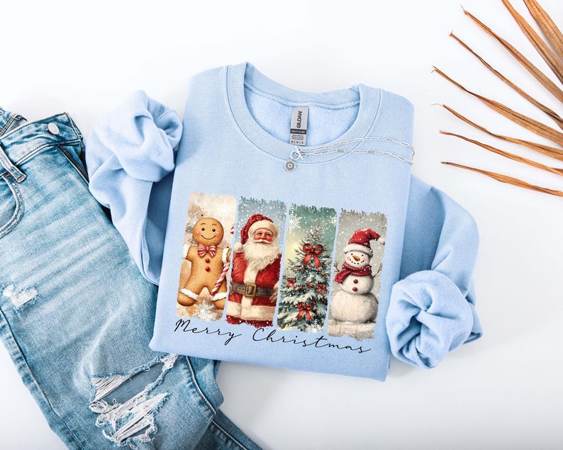 Puede incluir: Sudadera azul claro con un dise&ntilde;o navide&ntilde;o. El gr&aacute;fico incluye un hombre de jengibre, Pap&aacute; Noel, un &aacute;rbol de Navidad y un mu&ntilde;eco de nieve. Las palabras "Merry Christmas" est&aacute;n escritas debajo. La sudadera se muestra con unos vaqueros azules.