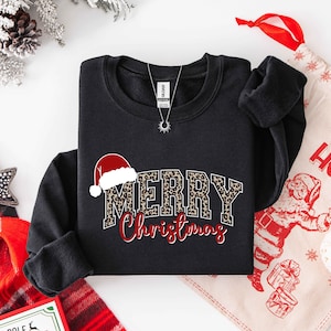 May include: Black sweatshirt with "MERRY Christmas" text. The word "MERRY" has a leopard print design, and the word "Christmas" is in red script. A Santa hat graphic is above the word "MERRY". A silver necklace is visible.