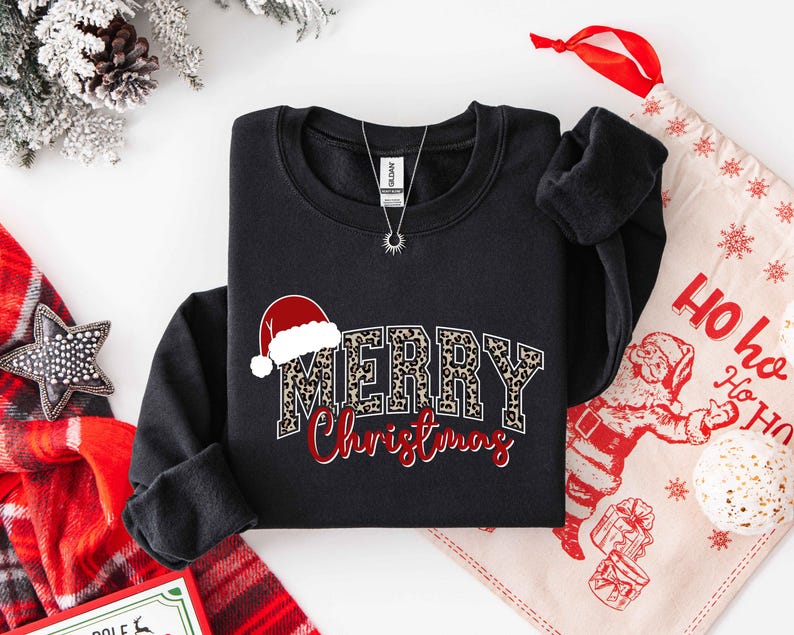 May include: Black sweatshirt with "MERRY Christmas" text. The word "MERRY" has a leopard print design, and the word "Christmas" is in red script. A Santa hat graphic is above the word "MERRY". A silver necklace is visible.