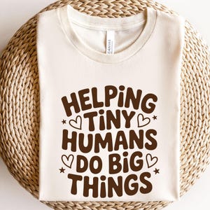 May include: Cream-colored t-shirt with the text "HELPING TINY HUMANS DO BIG THINGS" in brown, with heart and star accents. The shirt is made of a soft material and is suitable for everyday wear.