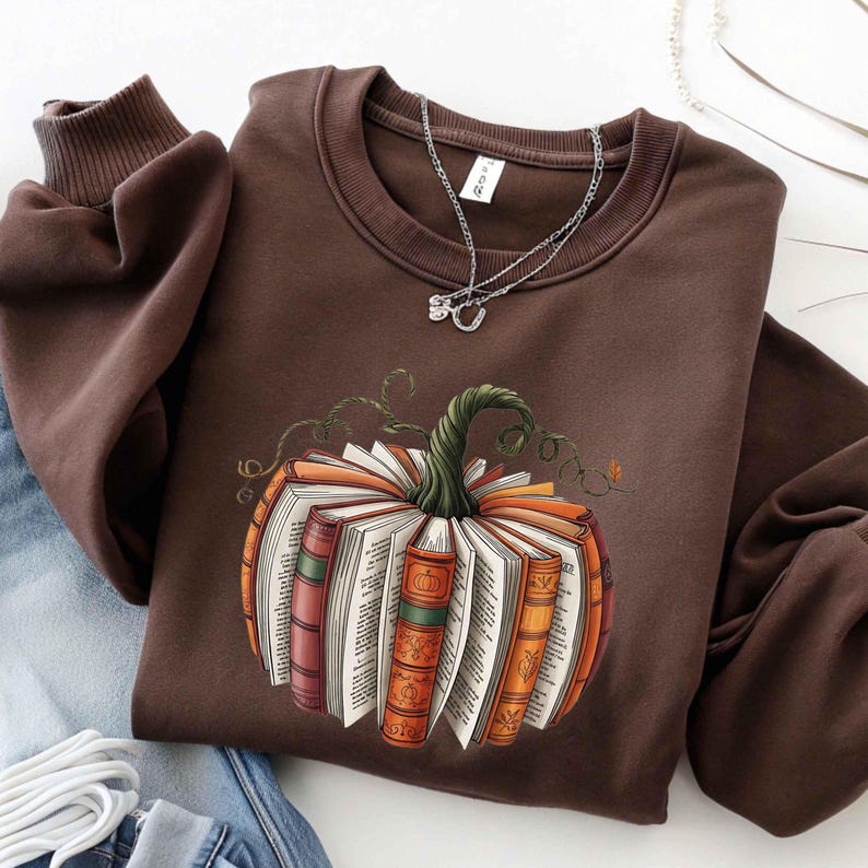 May include: A brown sweatshirt featuring a pumpkin design made of open books. The pumpkin has a green stem and vine details. The sweatshirt has a crew neck and long sleeves. A silver necklace is visible.