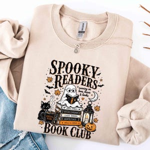 May include: A beige crewneck sweatshirt featuring a graphic print with the text "SPOOKY READERS BOOK CLUB" and "running on boos, brews & books". The design includes a ghost reading a book on a stack of books, a black cat, a lantern, and a pumpkin.