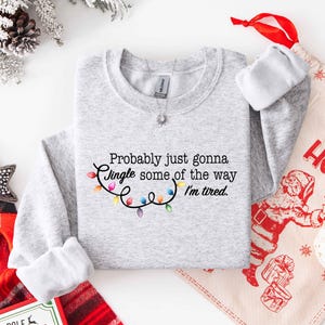 Puede incluir: Sudadera gris claro con la frase "Probably just gonna Jingle some of the way I'm tired." y una cadena de luces navide&ntilde;as de colores. Un tema festivo navide&ntilde;o.