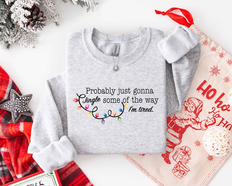 Puede incluir: Sudadera gris claro con la frase "Probably just gonna Jingle some of the way I'm tired." y una cadena de luces navide&ntilde;as de colores. Un tema festivo navide&ntilde;o.