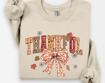 Könnte beinhalten: Beiges Sweatshirt mit dem Wort "THANKFUL" in einer Mischung aus Mustern und Farben. Die Buchstaben haben Karomuster, Leopardenmuster und florale Designs. Unter dem Wort befindet sich eine große Schleife mit Leopardenmuster, mit Herbstblättern und Blumen.