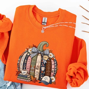 May include: A bright orange crewneck sweatshirt featuring a graphic of a pumpkin constructed from stacked books. The books have spines labeled with text including Gilmore Girls, Stars Hollow, and In Omnia Paratus, accented by a blue gingham bow and daisies.